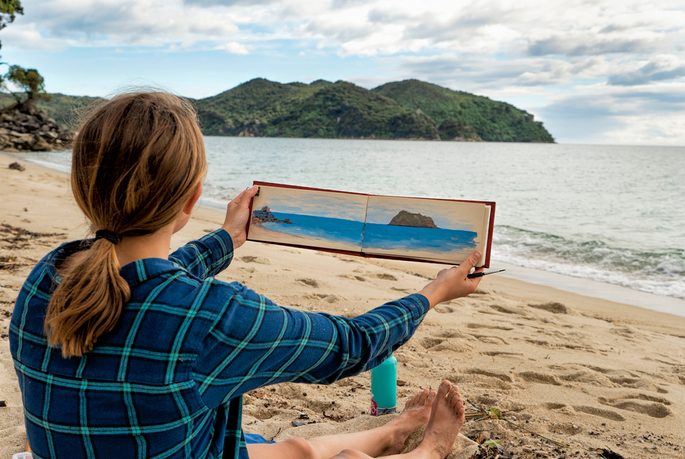 A woman holds a painting while sitting on a beach