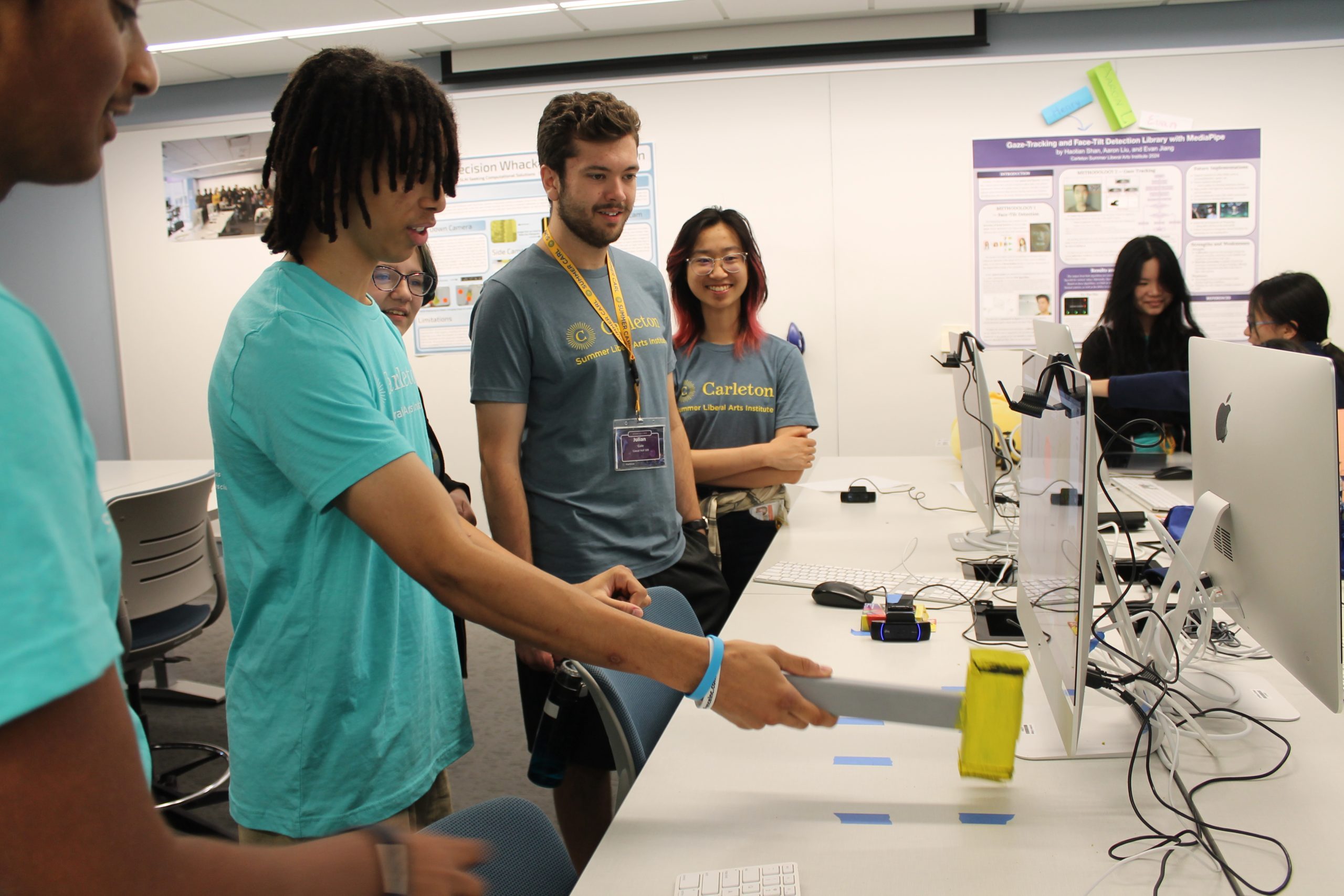 A student uses a mallot to play a computer science game created by another student while other students look on smiling.