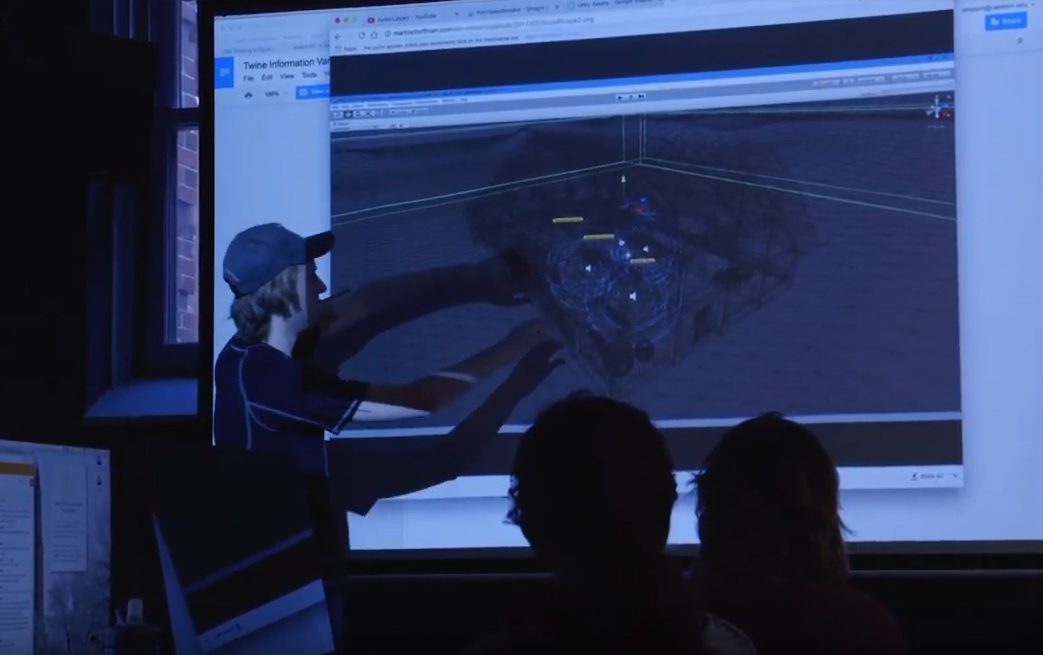 A student stands before a screen where a 3D digital image is projected