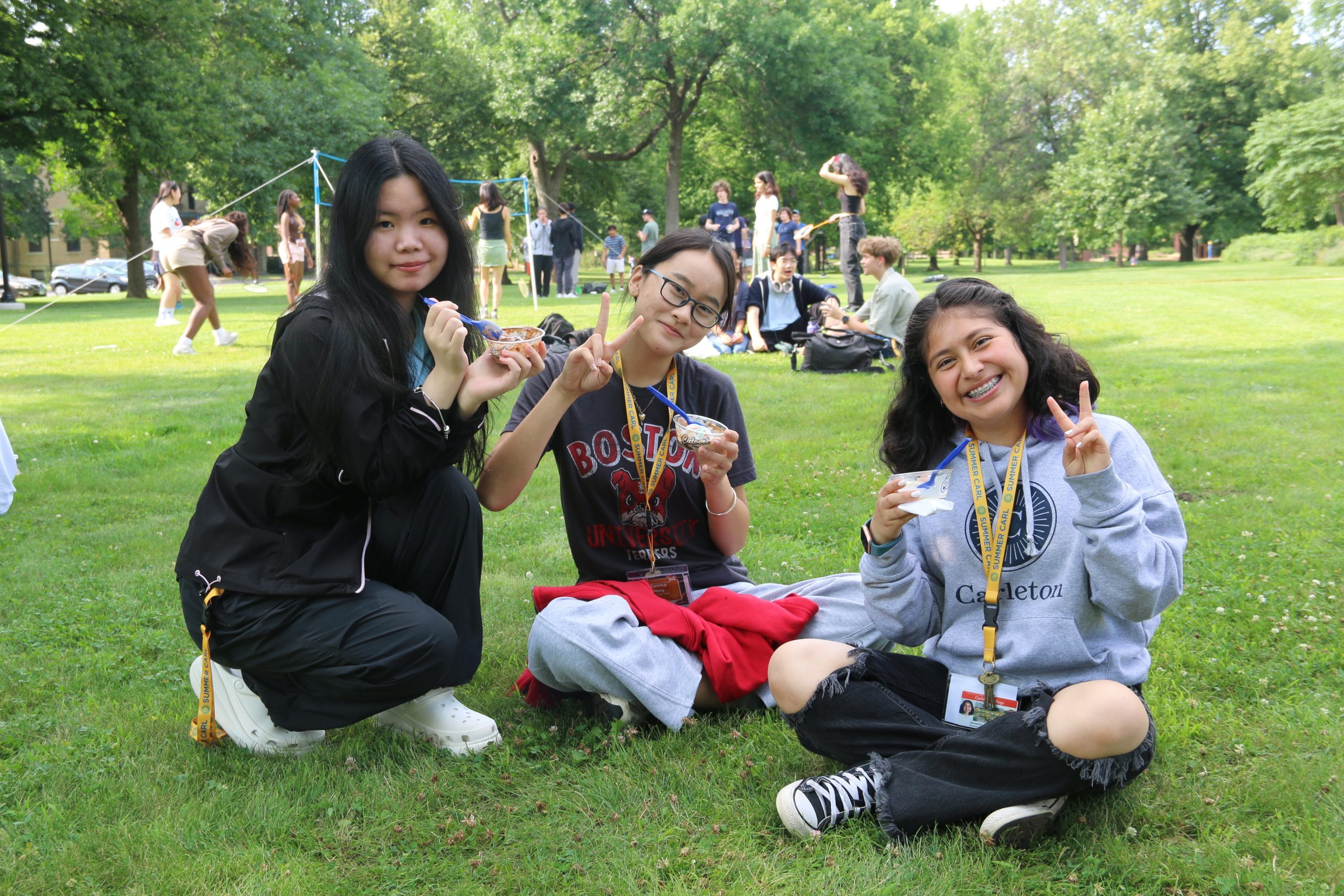 Three girls eat ice cream in the grass while groups of students sit in the grass and play volleyball behind them.
