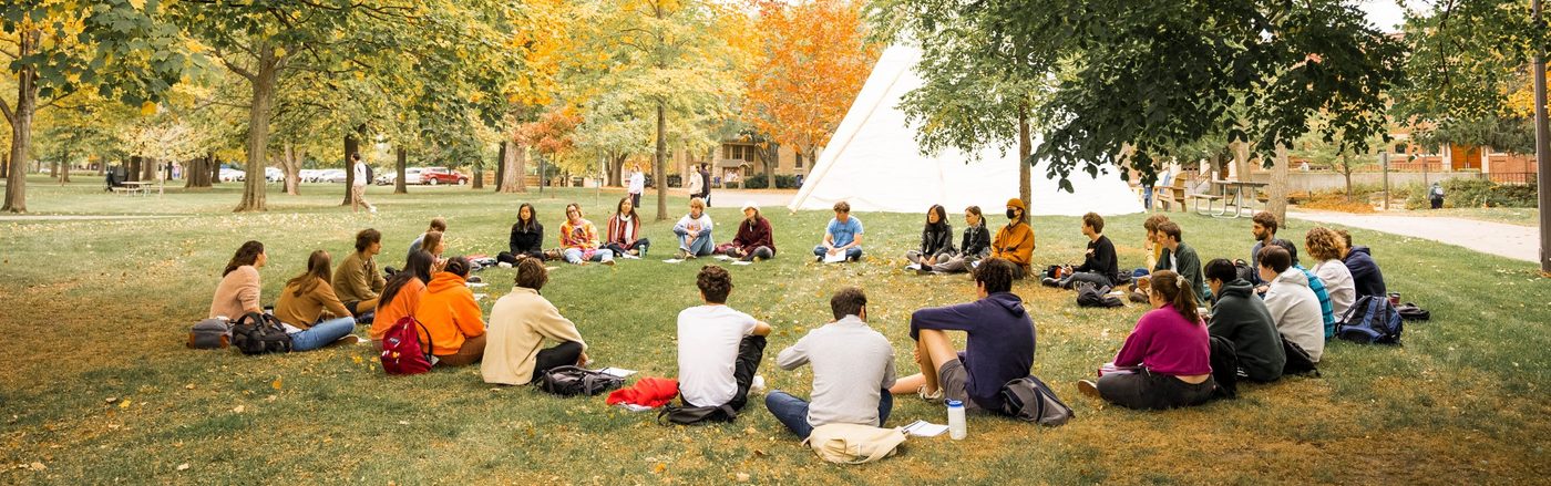 A class meets outside sitting in a circle, with a native American tipi in the background