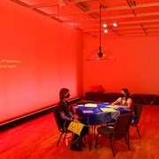 Two people sitting at a table in a room lit with red light, heating pads and cords are strewn about chairs and benches. On the wall is projected a transcription of an audio installation of Finnegan Shannon and Ezra Benus that addresses the culture around heating pads.