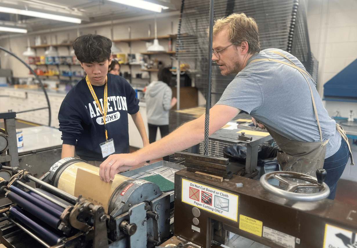 An instructor assist a student with an ink press in a studio art space.